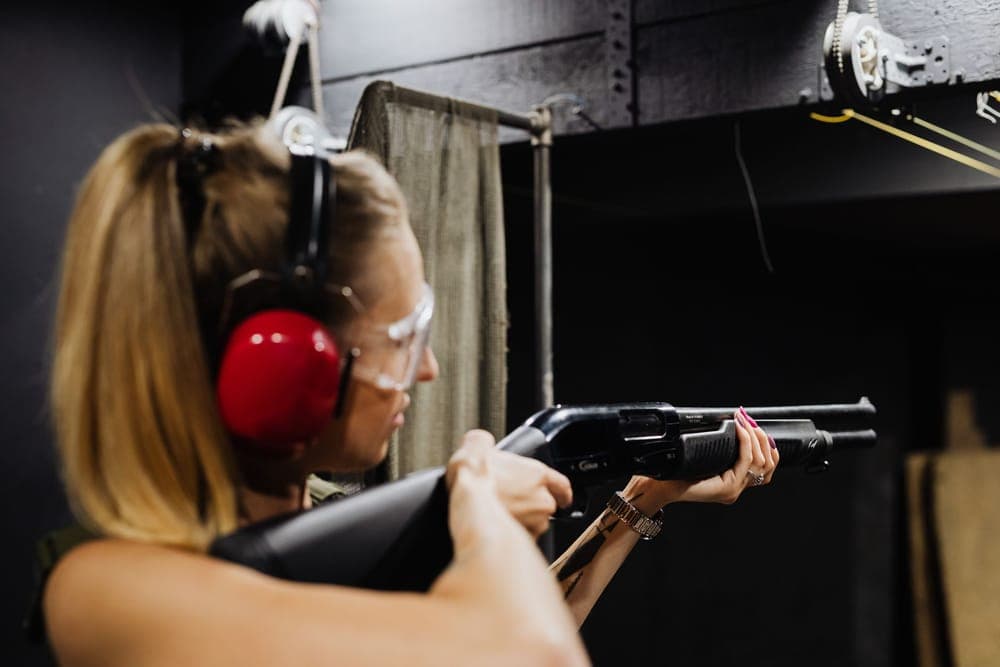 A woman in red earmuffs and clear safety glasses aims a shotgun at an indoor range