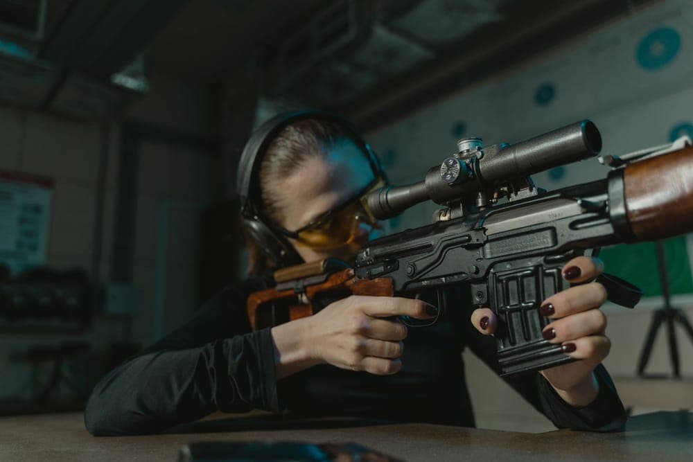A woman wearing earmuffs and amber glasses aims a scoped rifle at an indoor range