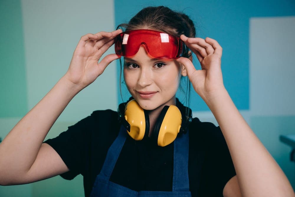 Illustrative stock photo: a smiling woman adjusting red safety glasses with yellow earmuffs around her neck