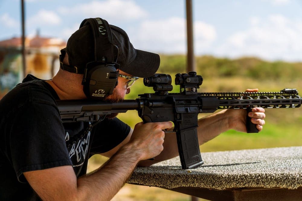 Illustrative stock photo: a shooter wearing electronic earmuffs and glasses aims from a bench at an outdoor range